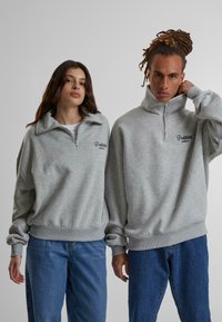 Sudaderas grises con cremallera y cuellos altos, con puños y dobladillos acanalados, combinadas con jeans azules. "Prendas Prohibidas" bordado en la parte frontal.