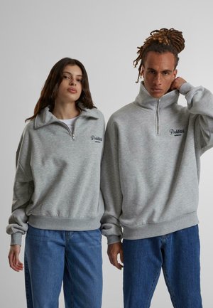 Sweatshirts zippés gris avec col haut, présentant des poignets et des ourlets côtelés, associés à un jean bleu. "Vêtements Interdits" brodé à l'avant.