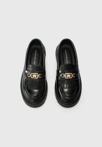 Par med svarte skinnloafers med tykke såler, dekorative gullfargede logospenner og frynse detaljer på hvit bakgrunn.