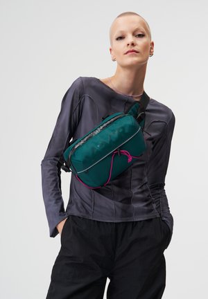 Person trägt eine türkisfarbene Umhängetasche mit schwarzen und pinken Akzenten über einem langärmligen grauen Oberteil und schwarzen Hosen vor einem schlichten Hintergrund.