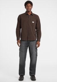 Camicia a maniche lunghe a quadri marrone con taschino sul petto, abbinata a pantaloni cargo grigi larghi con tasche laterali e scarpe nere.
