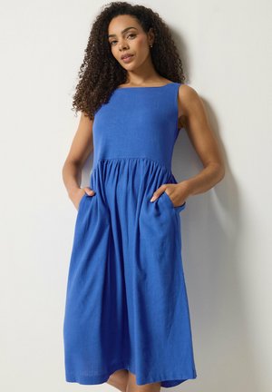 Frau mit lockigem Haar, die ein ärmelloses, königsblaues Kleid mittlerer Länge mit Taschen trägt und vor einer weißen Wand steht.