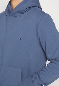 Felpa blu realizzata in tessuto morbido con tasca a marsupio, caratterizzata da un piccolo logo bianco, rosso e blu sul petto. Maniche lunghe con polsini a costine.