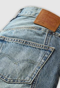 Jeans di denim azzurri chiari con una patta in pelle marrone, caratterizzati da cuciture visibili e un design classico della tasca posteriore.