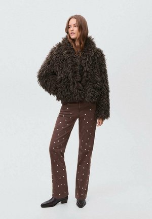 Donna che indossa una giacca fluffy marrone scuro, pantaloni marroni con pois bianchi e stivaletti neri alla caviglia, in piedi contro uno sfondo semplice.