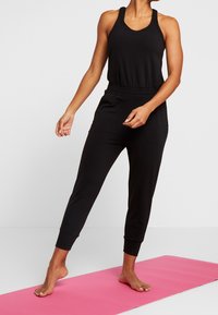 Un débardeur noir sans manches et un pantalon de jogging assorti, en matériau doux avec une coupe décontractée, accompagnés d'un tapis de yoga rose.