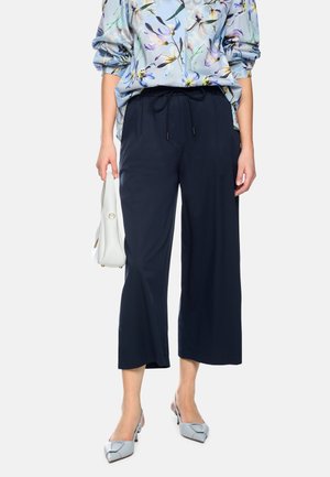 Woman wearing wide-leg navy pants with drawstring waist, light blue floral blouse, silver pointed-toe heels, and holding a white handbag.