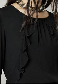 Blusa nera con colletto arricciato e maniche lunghe. Al collo è indossato un collier in argento spesso, che aggiunge un tocco metallico.