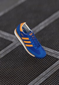 Blue and orange athletic shoe featuring a suede upper, three stripes design, and a white rubber sole. The laces are blue and textured.