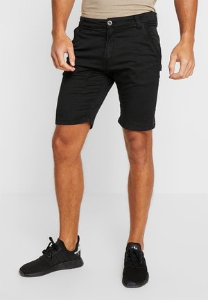 Mand iført sorte knælange shorts og sorte sneakers, stående mod en ensfarvet lys baggrund.