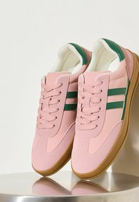 Rozā sporta apavi ar divām zaļām svītrām, izgatavoti no gludiem un fakturētiem materiāliem, ar gumijas zoli un apaļu purngalu.