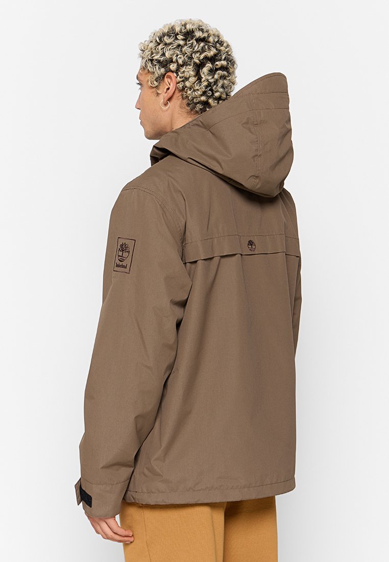 Brązowa ocieplana kurtka z kapturem, z poziomym szwem na plecach i naszywką z logo na rękawie. Materiał wydaje się gładki i wodoodporny.