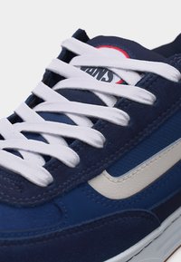 Κοντινό πλάνο αθλητικού παπουτσιού σε μπλε ναυτικό χρώμα με λευκά κορδόνια, λευκή πλαϊνή ρίγα και στρογγυλό λογότυπο στη γλώσσα.