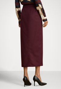 Burgunderfarget midilengde skjørt laget av glatt stoff, med en tettsittende midjebånd. Kombinert med svarte stiletthæler og mønstrede lange ermer.