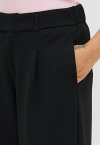 Calças pretas com um corte ajustado, apresentando uma textura suave, bolsos laterais e um ligeiro pregueado. A cintura é elástica para maior conforto.