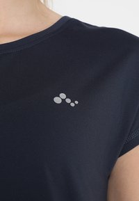 Detailní záběr na námořnickou modrou košili, která na hrudi ukazuje malou reflexní ikonu se pěti oválnými body uspořádanými v diagonální linii.