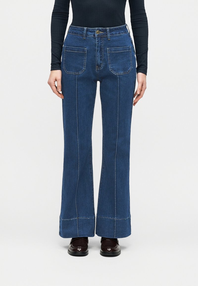 Άτομο που φοράει μπλε τζιν ψηλού μέσης με εμπρόσθιες τσέπες, καμπάνα στα πόδια, μαύρο μακρυμάνικο τοπ και σκούρα δερμάτινα παπούτσια.