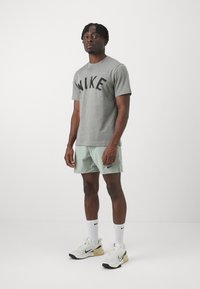 Pelēks īsām piedurknēm T-krekls ar tumšiem burtiem "NIKE", kombinēts ar gaiši zaļām šortiem un baltām sporta apavām ar melniem akcentiem.