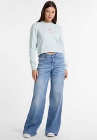 Lichtblauwe cropped sweatshirt met logo; wijde medium wash jeans en witte sneakers. De outfit heeft een relaxte pasvorm en een strakke uitstraling.