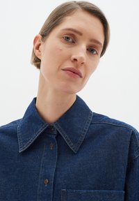 Dark blue denim shirt with a classic collar, button placket, and front pocket. The fabric has visible texture and subtle stitching details.