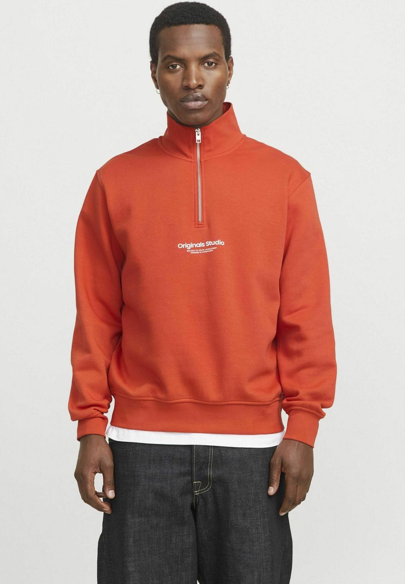 Sudadera naranja con cremallera y cuello alto, con el texto blanco "Originals Studio" en el pecho. Tejido suave y puños acanalados. Jeans oscuros.