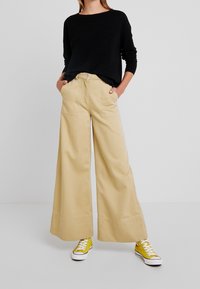 Beige vida byxor med framfickor och synliga sömdetaljer, kombinerat med en svart ribbad långärmad topp och gula sneakers.