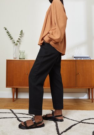 Person iført løs brun langærmet top, sorte kortere bukser og sorte sandaler med spænde, står på et mønstret tæppe indendørs.