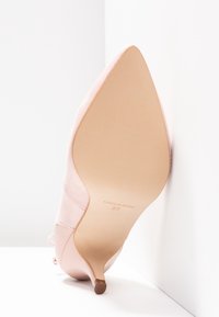 Chaussure à talon haut en daim rose à bout pointu, talon fin, semelle marquée "Made in Spain" et taille 37, positionnée verticalement contre un coin blanc.