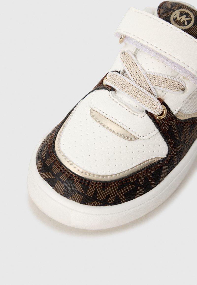 Baskets marron et blanches avec un bout perforé, des accents dorés et un logo. Elles disposent de lacets en tissu et d'une sangle Velcro pour la fermeture.