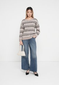 Gruba dziergana sweter w zygzakowy wzór w beżowych i szarych odcieniach, w połączeniu z szerokimi dżinsami w niebieskim kolorze i małą torebką w kolorze kremowym. Czarne buty na obcasie.