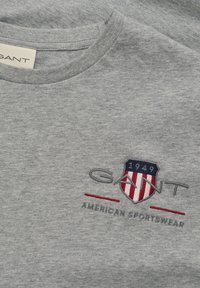 Pilkas trumpas rankovėmis marškinėliai, pagaminti iš medvilnės, su apvalia iškirpte ir siuvinėtu logotipu su skydu, juostelėmis ir užrašu.