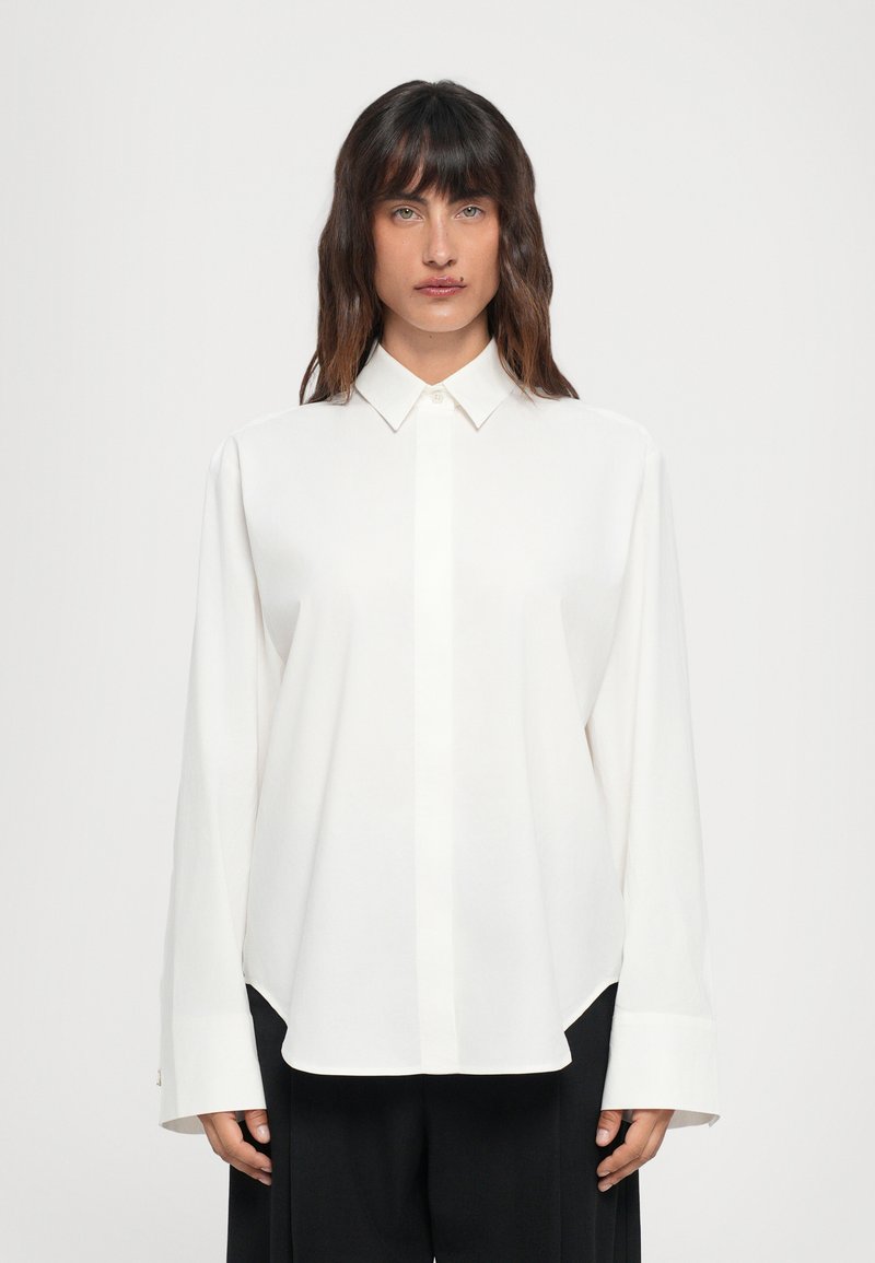 Camicia bianca a maniche lunghe con colletto classico, texture liscia e orlo leggermente arrotondato, abbinata a pantaloni ampi scuri.
