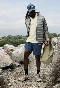 Grijze geïsoleerde jas, lichtbeige t-shirt, blauwe shorts, tan wandelschoenen, met een groene rugzak, op rotsachtig terrein.