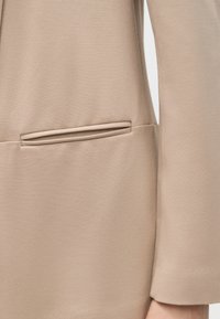 Κοντινό πλάνο μανικιού μπεζ σακακιού και τσέπης με καπάκι σε φόντο από λείο ύφασμα.