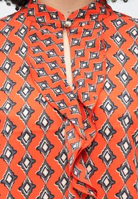 Sarkanā blūze ar dimanta rakstu tumši zilā un baltā krāsā, ar atslēgas formas izgriezumu pie kakla un volānu detaļām priekšpusē. Gluda auduma tekstūra.