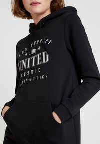 Černá mikina s kapucí vyrobená z měkké tkaniny, obsahující přední kapsu, stahovací kapucu a design s bílým textem včetně "Los Angeles," "United" a "Cosmic Aeronautics."