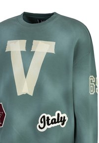 Der grüne Sweatshirt hat ein großes, beiges "V" mit Tape-Akzenten, "Italy" in weißer Stickerei und eine gemusterte "63" auf dem Ärmel.