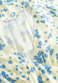 Blouse fleurie avec un col en V, ornée de petites fleurs bleues sur un fond jaune clair, fabriquée en tissu doux légèrement texturé.
