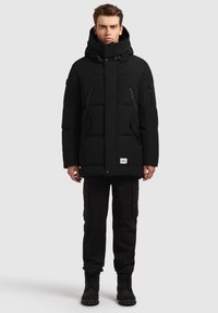 Juoda paminkštinta striukė su aukšta apykakle, kišenėmis su užtrauktukais ir spaudėmis užsegimu. Dėvima su juodomis kargo kelnėmis ir juodais batais. Paprastas dizainas.