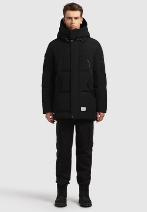 Svart vadderad jacka med hög krage, dragkedjefickor och tryckknappsstängning. Bärs med svarta cargobyxor och svarta stövlar. Enkel design.