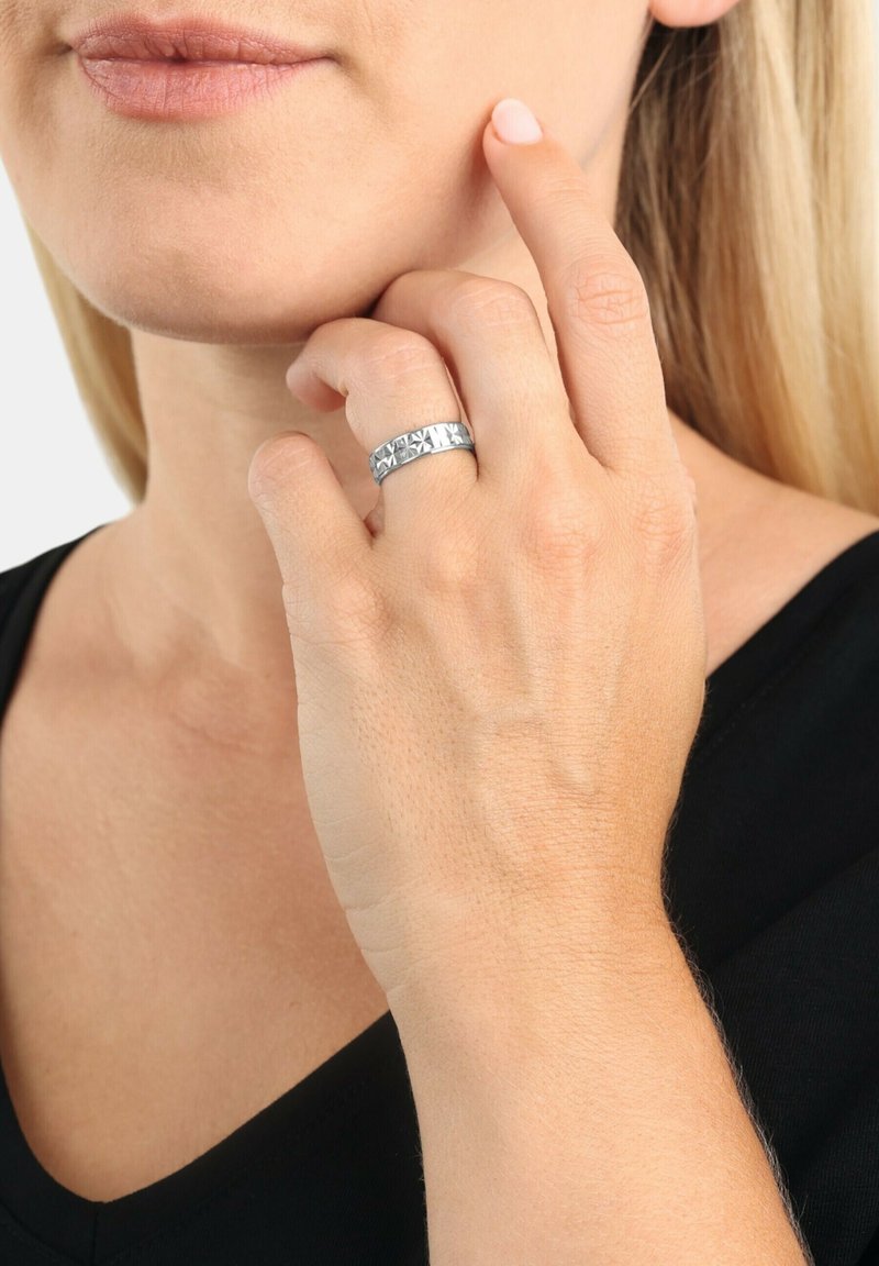 Anello in argento con motivo geometrico, finitura lucida e accenti opachi, indossato su un dito, mano poggiata sul mento.