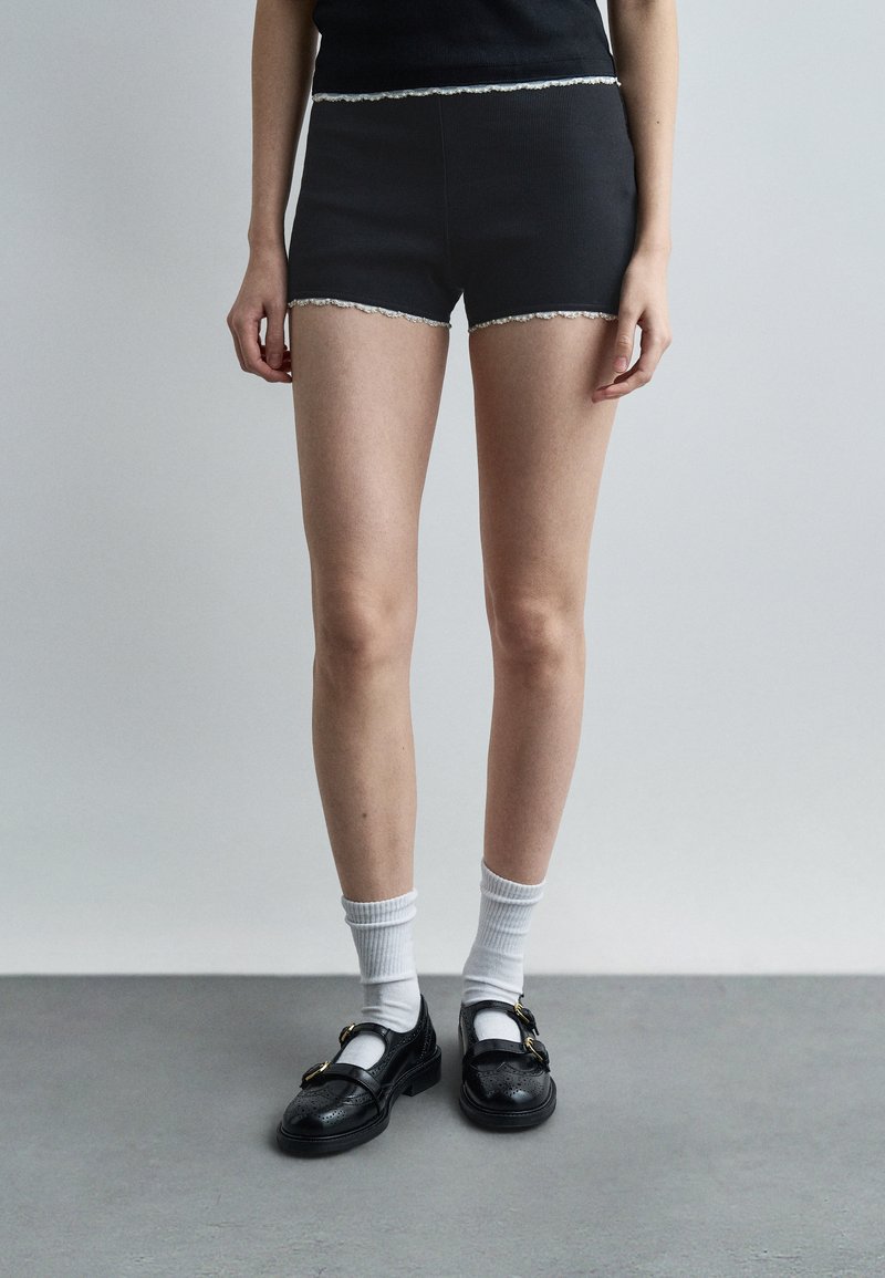 Personne portant un short côtelé noir avec bordure blanche, des chaussettes blanches montantes et des chaussures noires à boucle, debout devant un fond gris uni.