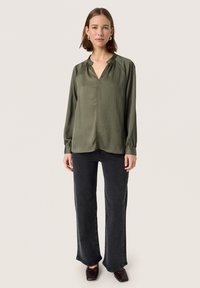 Blusa plissettata verde oliva con scollo a V, maniche lunghe e polsini arricciati. Abbinata a jeans neri a zampa d'elefante e scarpe marroni.