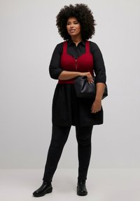 Red structured top with thin straps over a black button-up dress. Black skinny pants and ankle boots complete the outfit. Holds a black clutch bag.