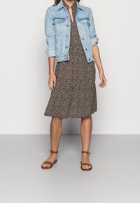 Jeansjacka över en svart klänning med geometriskt mönster, knälång, utsvängd nederkant, i kombination med bruna platta sandaler.