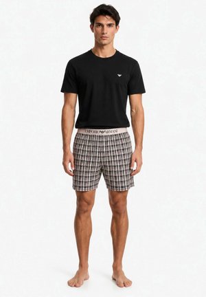 Uomo in piedi a piedi nudi, indossa una maglietta nera a maniche corte e pantaloncini a quadri con logo Emporio Armani sulla vita e sul petto.