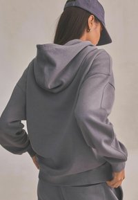 Szara bluza z kapturem wykonana z miękkiego materiału, w luźnym kroju, z ściągaczami przy mankietach i dolnej krawędzi oraz z kieszenią kangurzą z przodu.