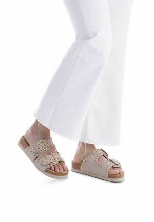 Sandalias tipo slide de ante beige con correas de doble hebilla y detalles con tachuelas, montadas sobre una suela ligera texturizada. Se llevan con pantalones blancos acampanados.