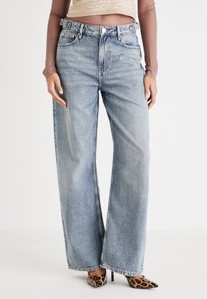 Kvinde iført højttaljede, vide, falmede blå jeans, gennemsigtig bluse med lange ærmer og leopardmønstrede, spidse høje hæle, står på hvidt gulv.