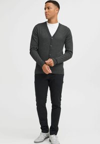 Mørkegrå cardigan med V-hals, knaplukning og lange ærmer, parret med sorte bukser og grå sneakers foran en neutral baggrund.